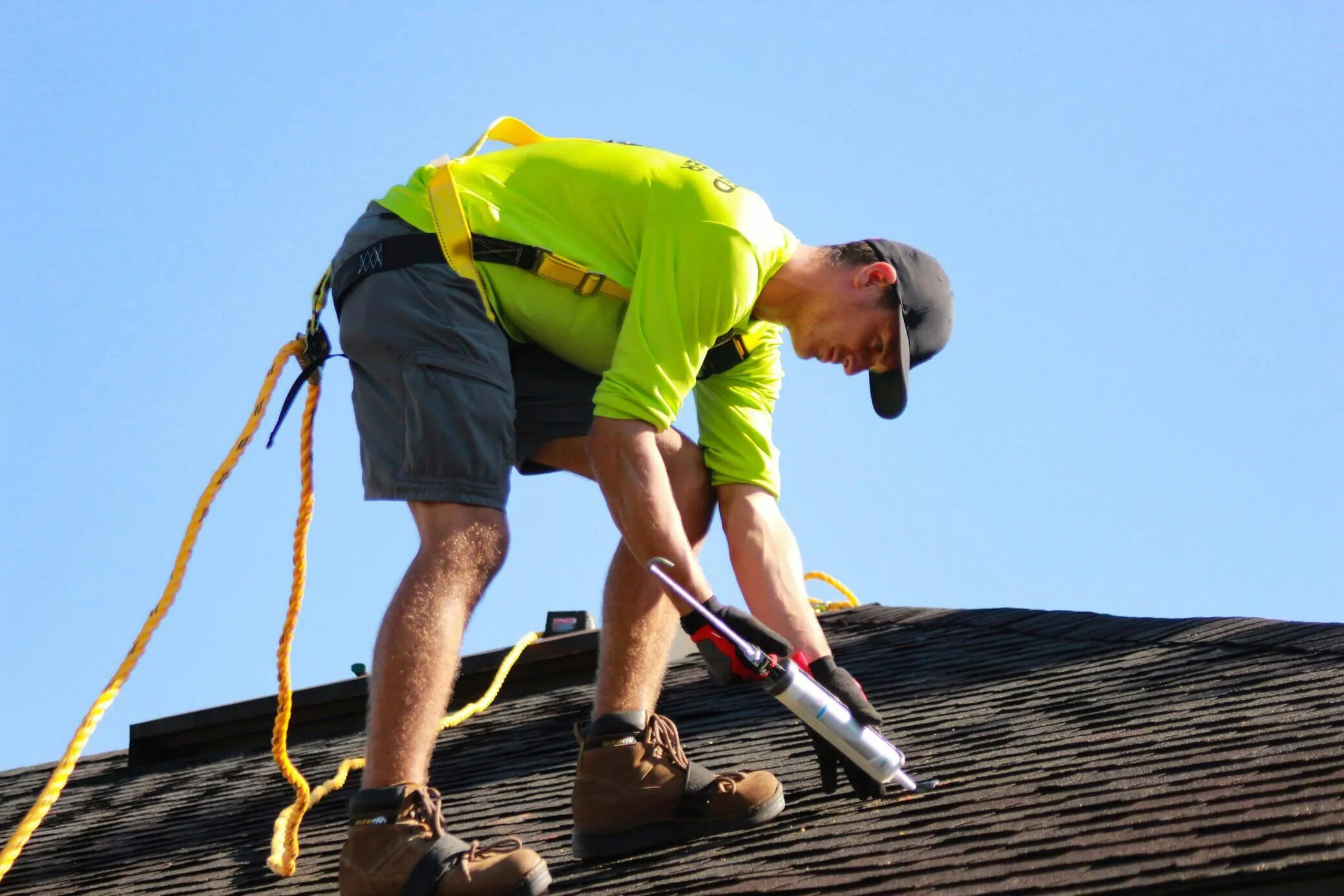 Roofer applying sealant on roof shingles while wearing a safety harness